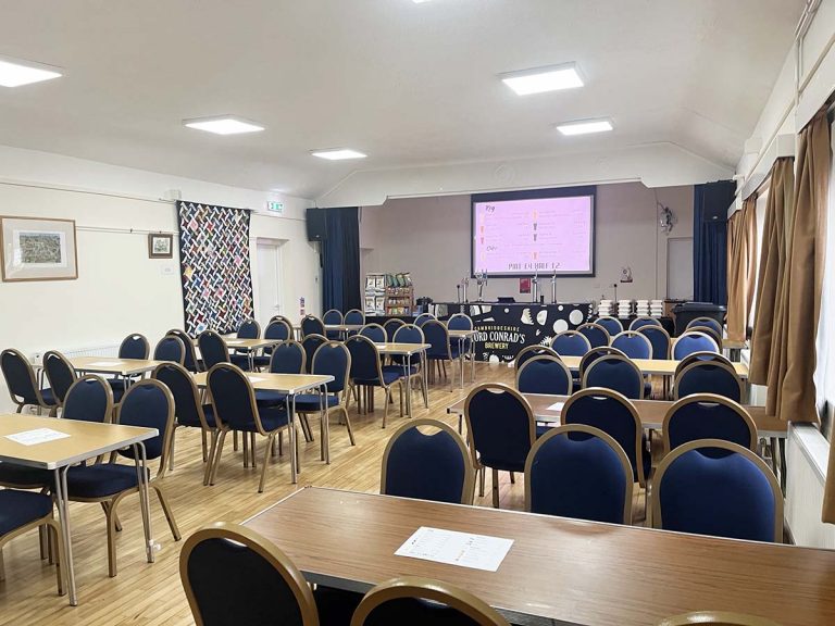 The main hall is shown with tables set out for a meal. At the back a bar can be seen with a video screen above it.