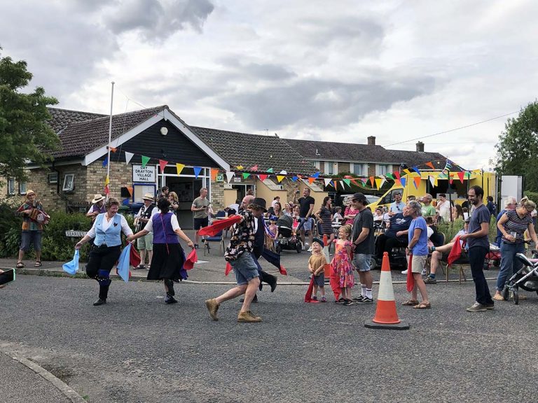 Morris Dancers perform in front of a crowd. In the background, crowds gather to order from a food van. The village hall at the back is decorated with bunting,